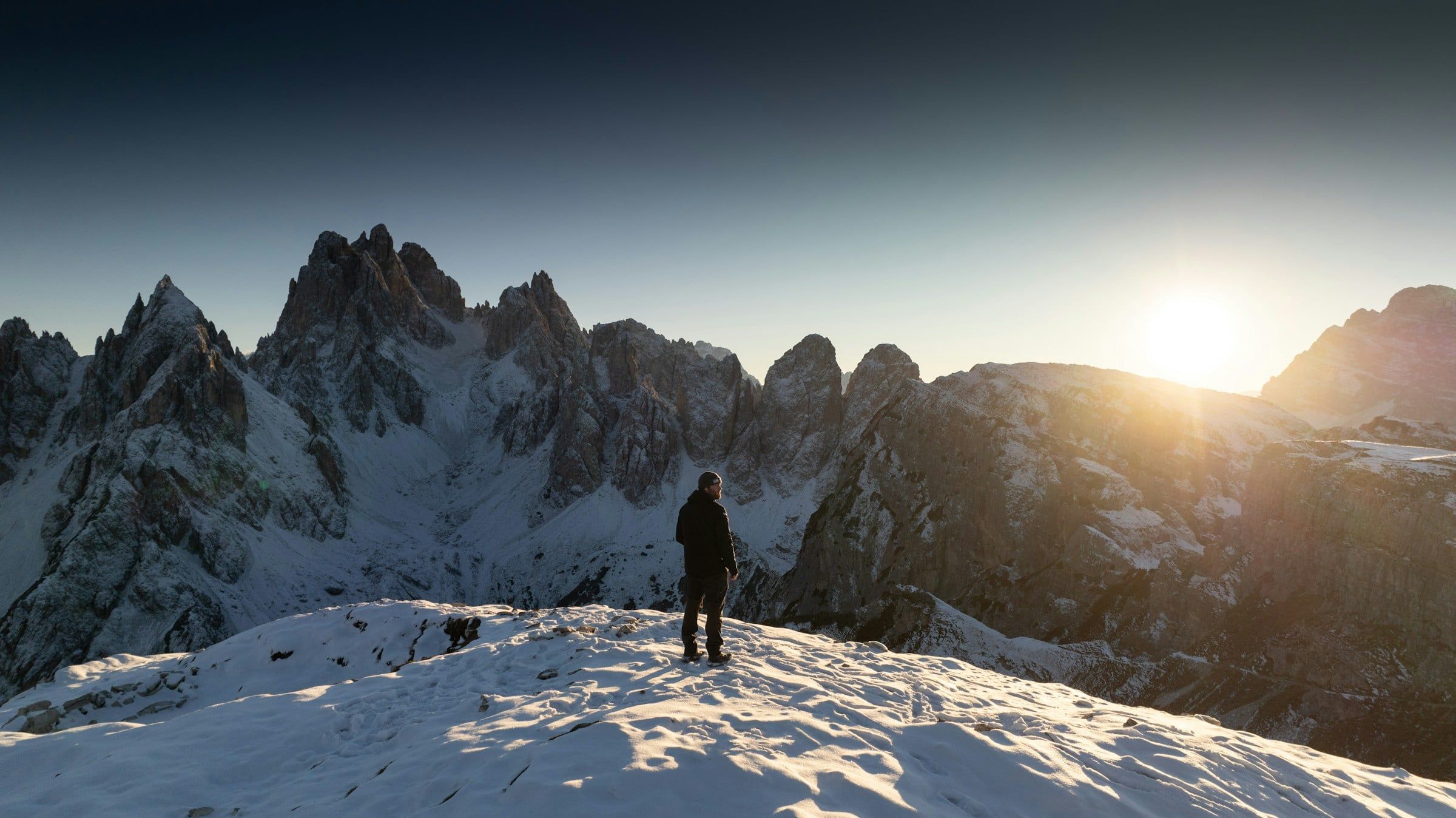 Alpinista in controluce su una cresta innevata, davanti a guglie rocciose illuminate dal sole basso che spunta tra le cime