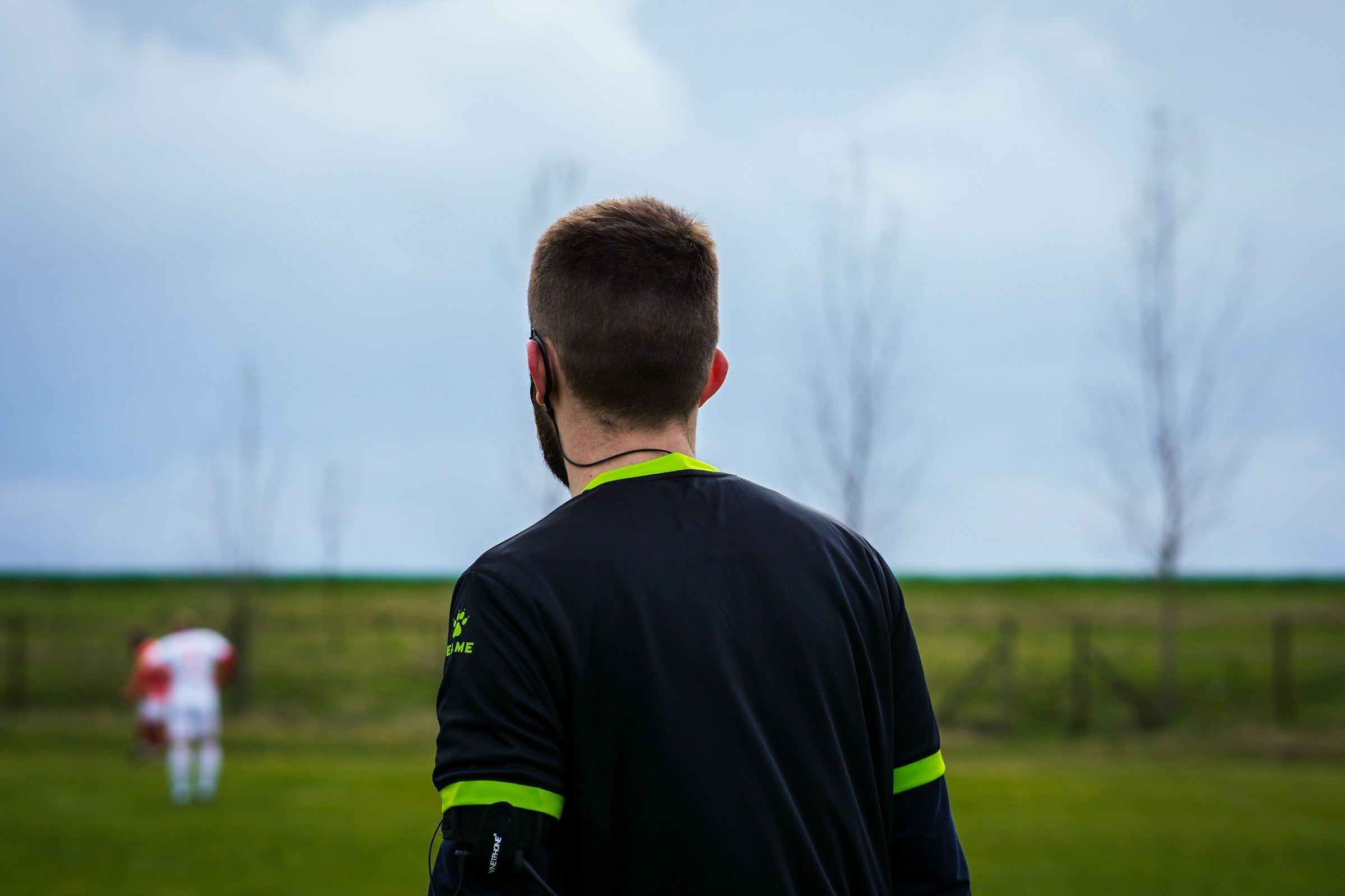 Un arbitro ripreso di spalle a bordo campo osserva una partita di calcio su un prato verde, sotto un cielo grigio e nuvoloso