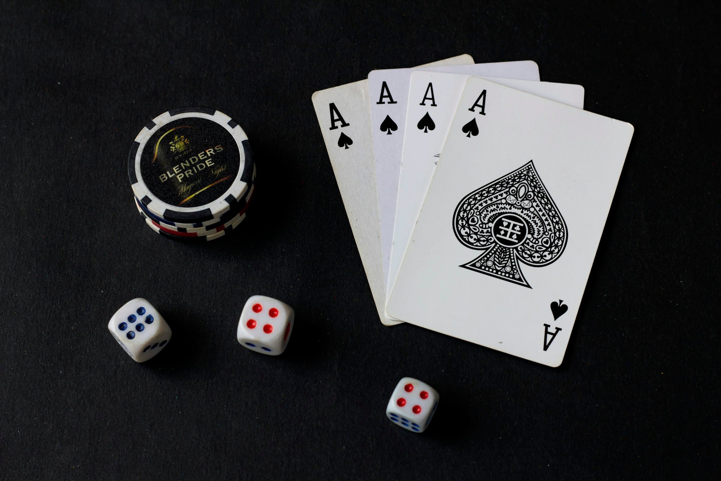 Quattro assi da gioco, tre dadi bianchi e una fiche da poker disposti su un fondo nero, illuminati dall'alto