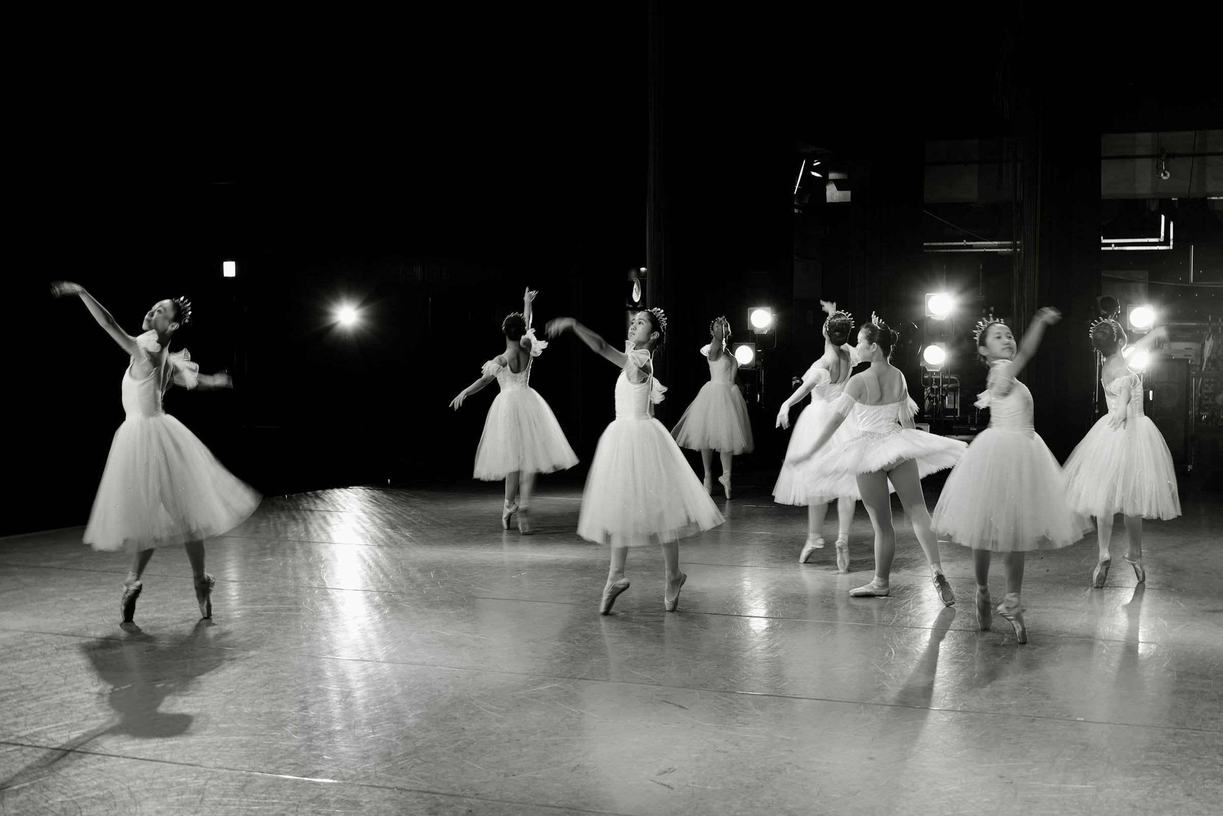 Ballerine in tutù bianco danzano sul palco sotto le luci di scena, in una foto in bianco e nero con i movimenti leggermente mossi