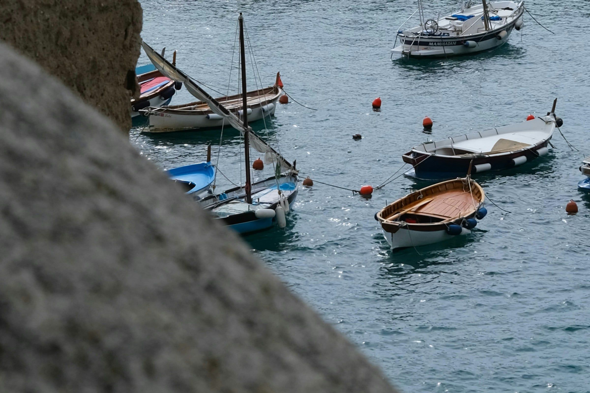 Piccole barche ormeggiate nell'acqua azzurra e trasparente di un porticciolo, viste dall'alto oltre il bordo di uno scoglio