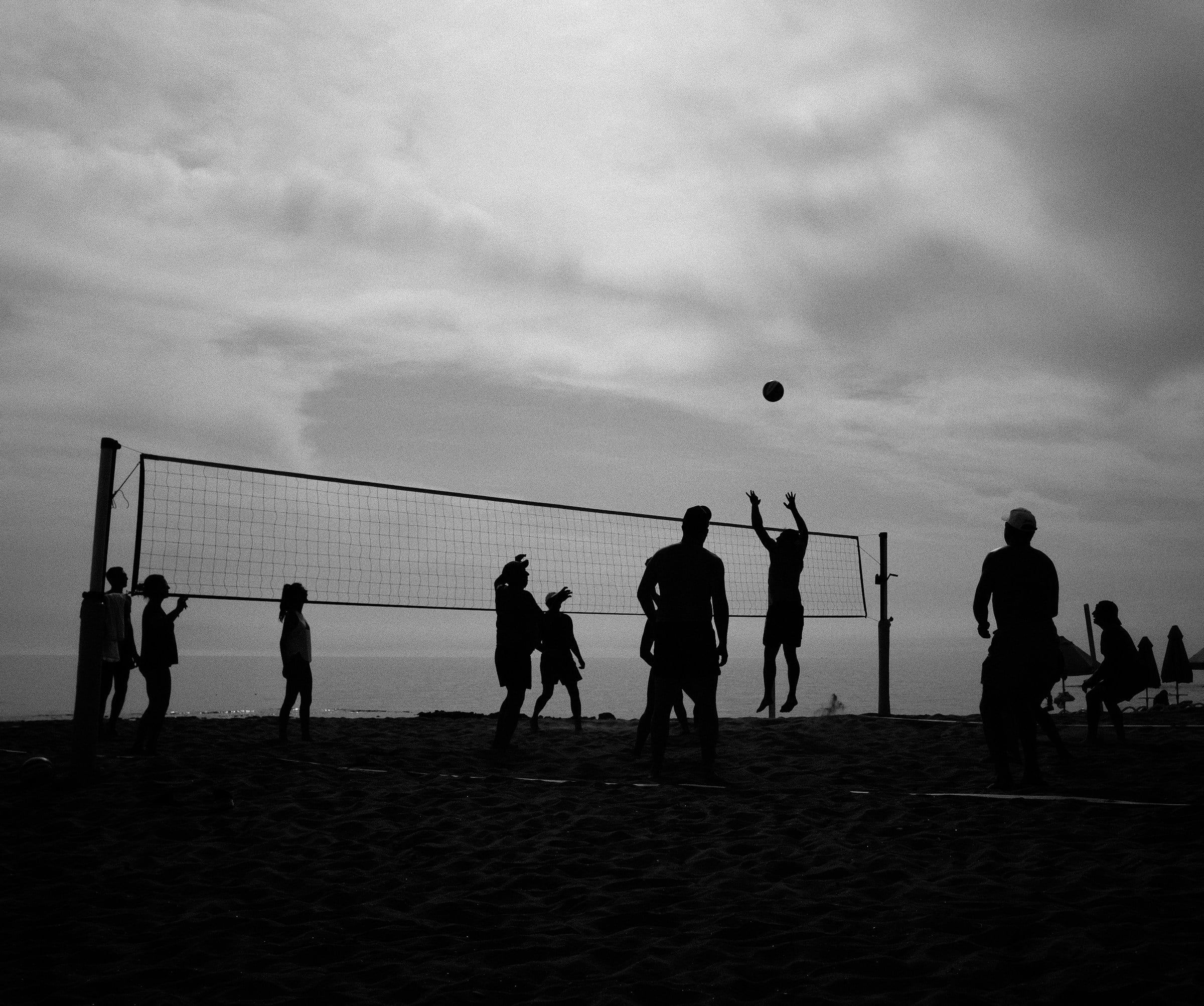 Silhouette di giocatori di beach volley in controluce sulla spiaggia, con la rete tesa e la palla sospesa in aria sopra di loro