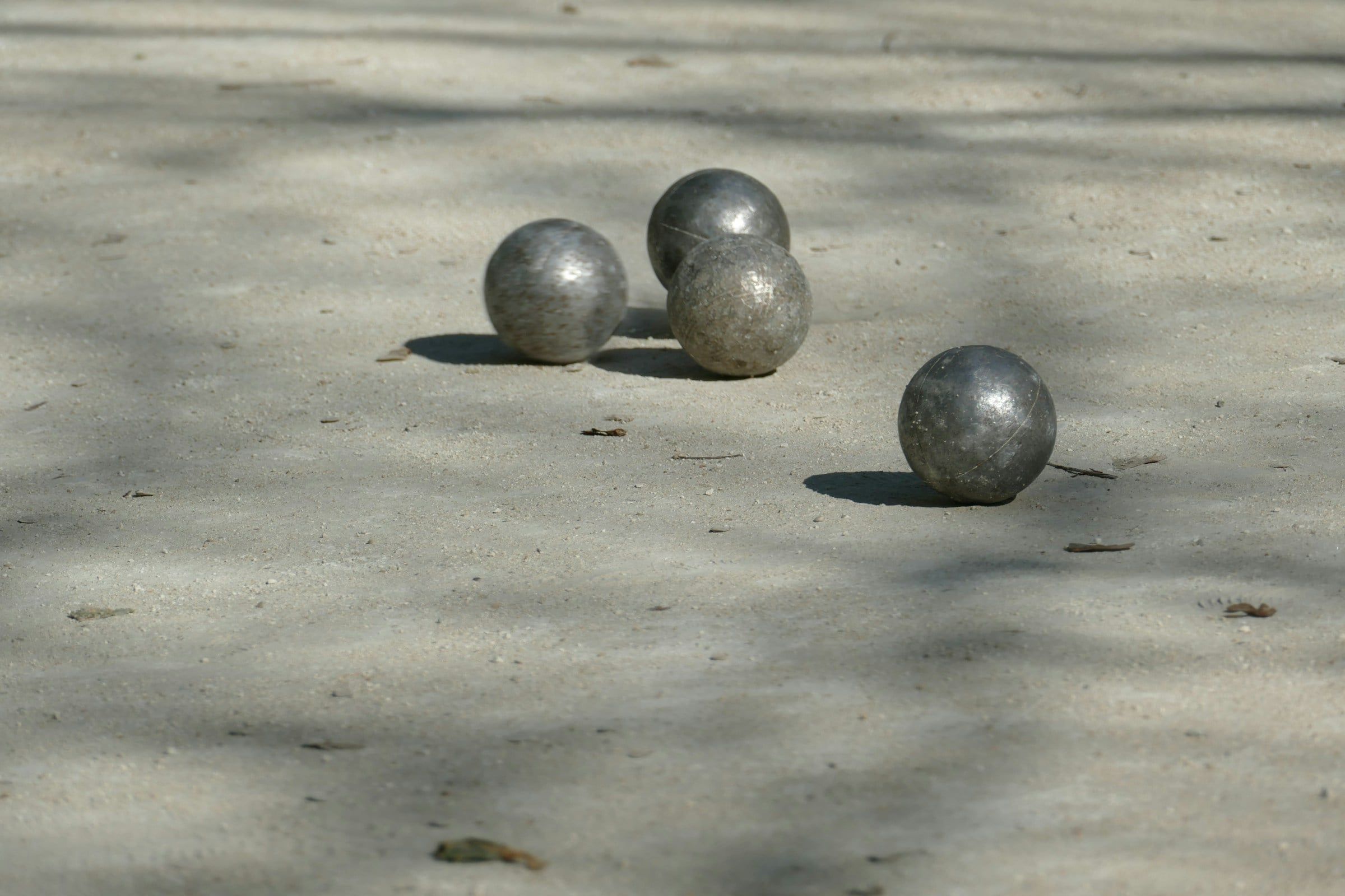 Quattro bocce in acciaio ferme su un campo di terra battuta chiara, con le ombre degli alberi che si allungano sul terreno