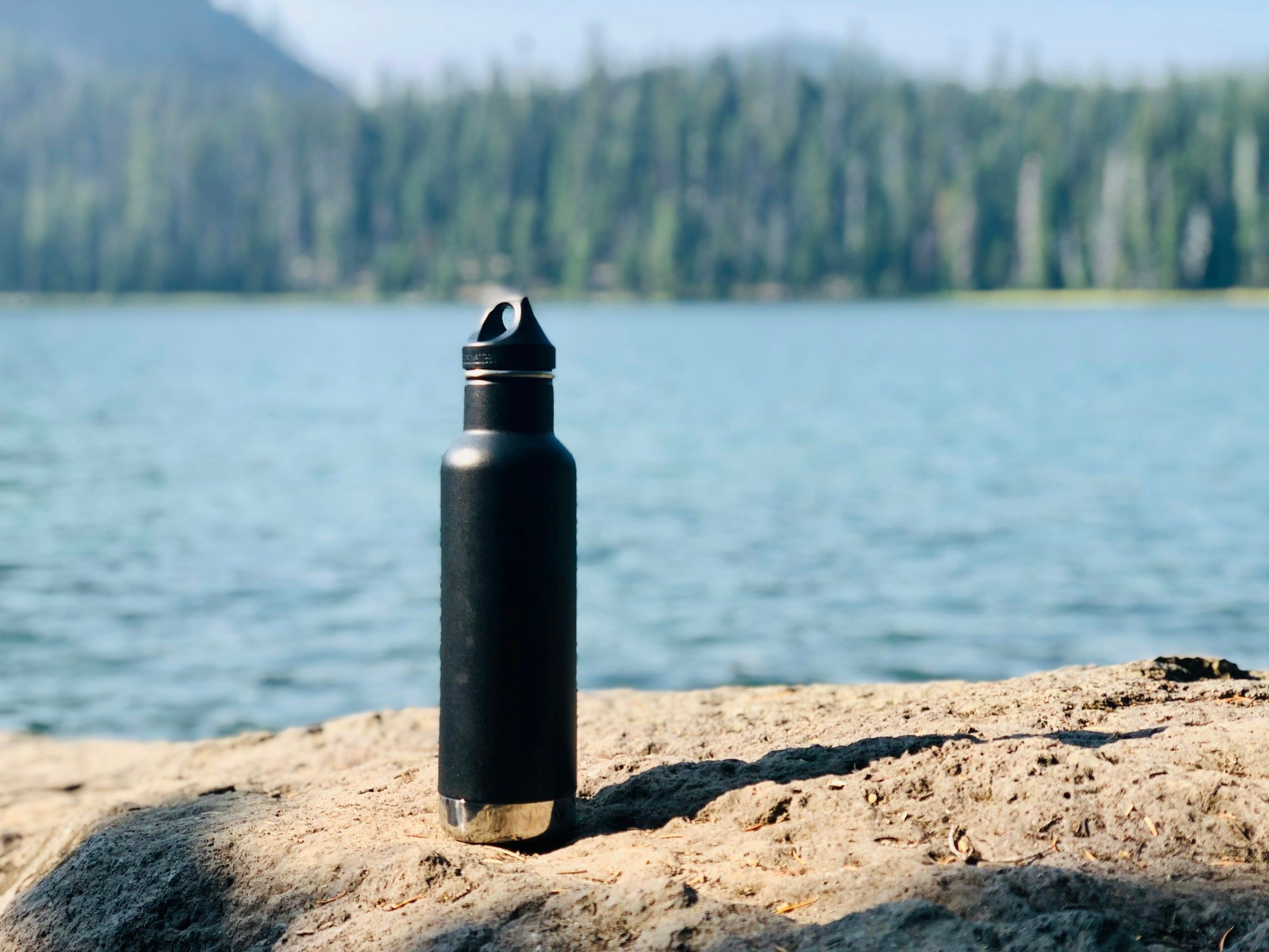 Borraccia termica nera appoggiata su una roccia in riva a un lago di montagna, con il bosco di abeti sullo sfondo