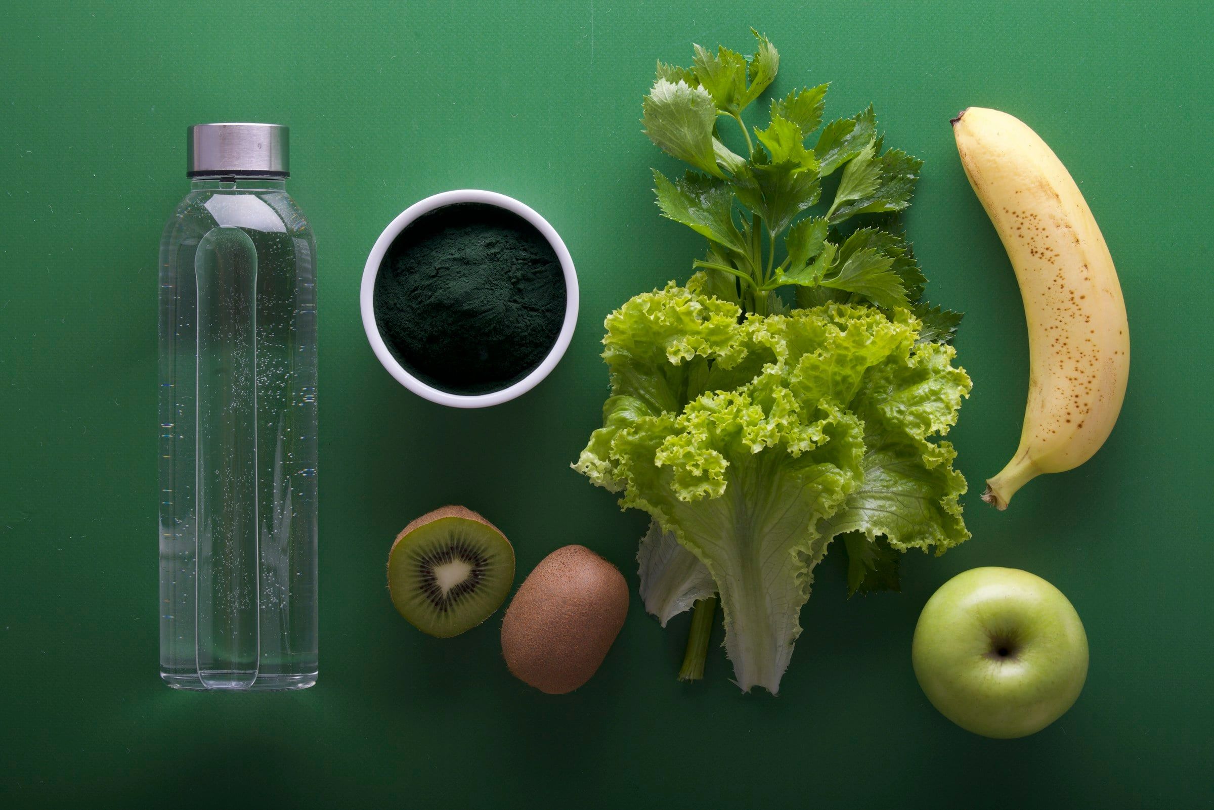 Bottiglia d'acqua in vetro accanto a una ciotola di spirulina, insalata, una banana, un kiwi e una mela verde su un fondo verde