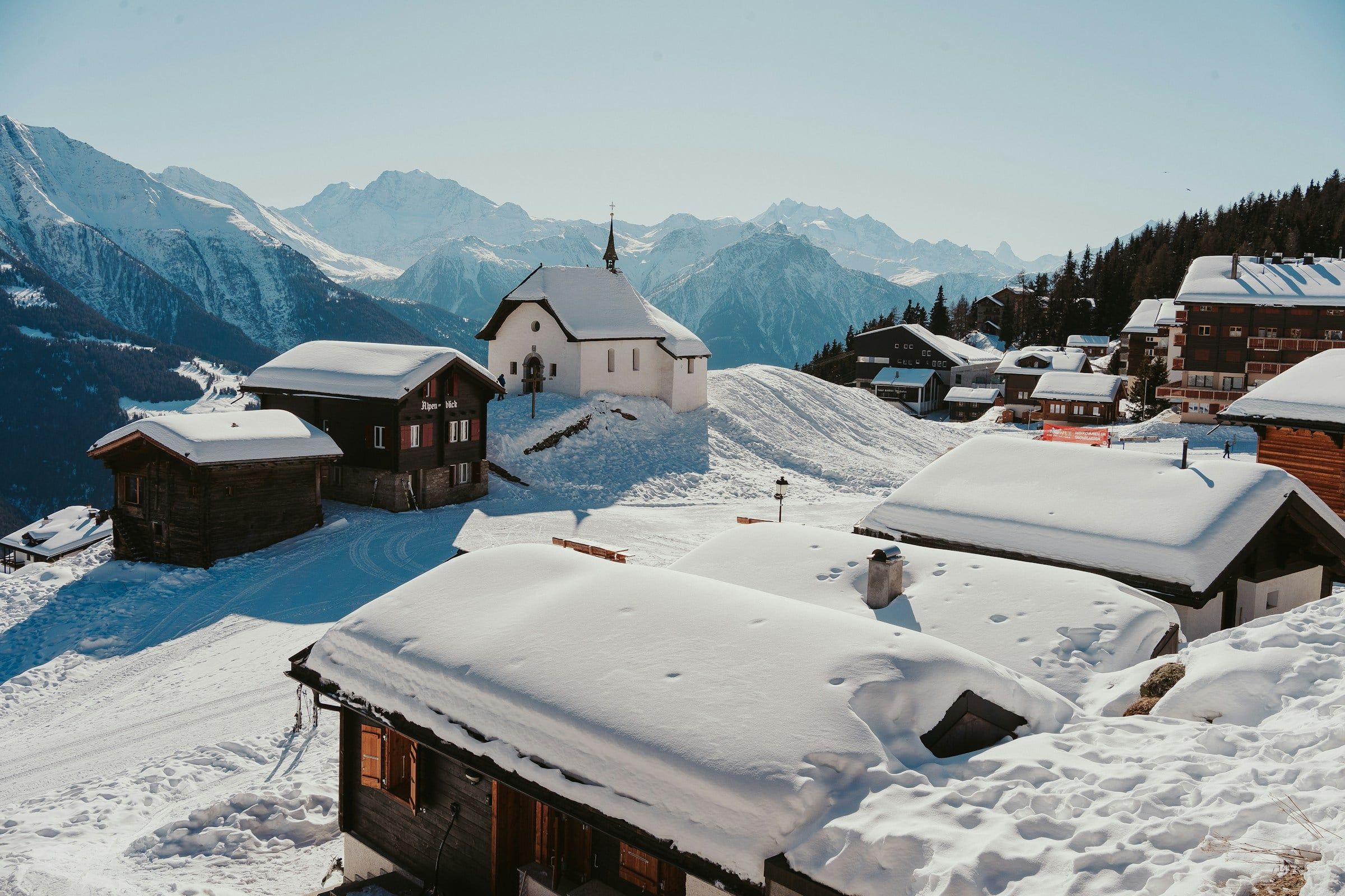 Chalet in legno coperti di neve in un villaggio di montagna, con una piccola chiesa bianca e le cime alpine sullo sfondo