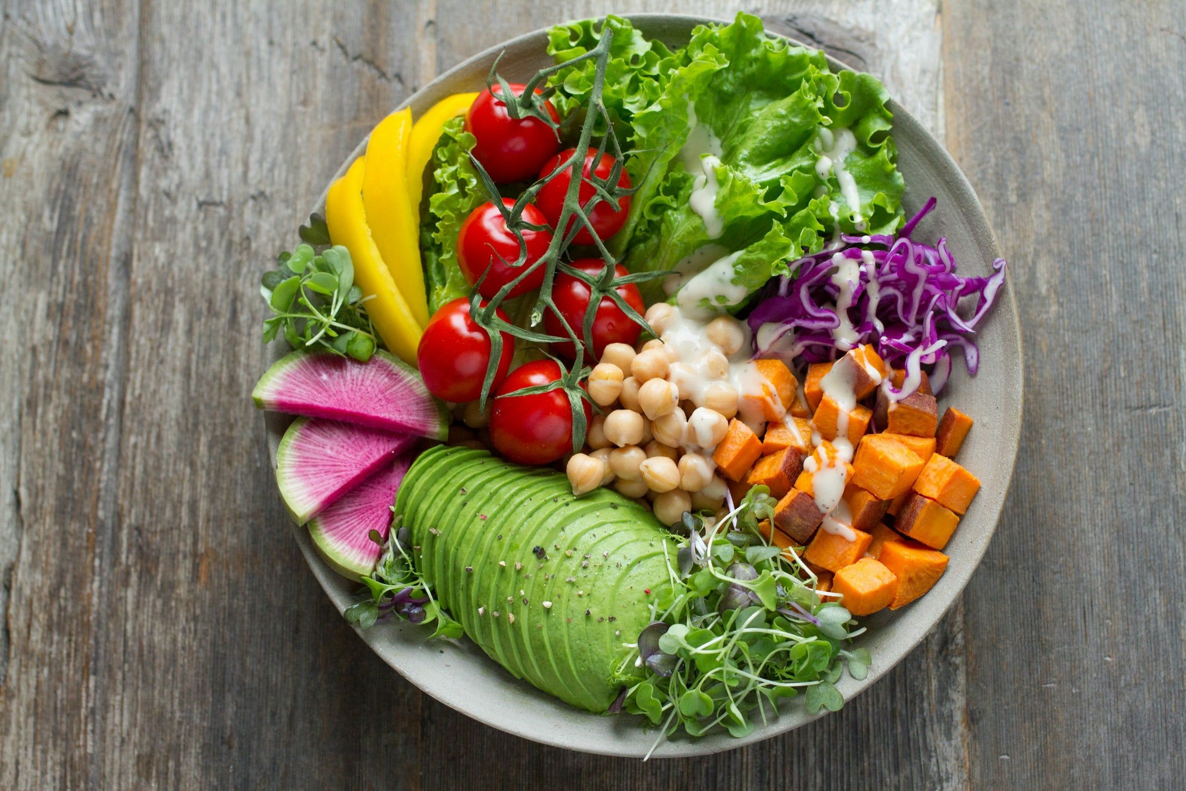 Ciotola di insalata colorata con avocado, pomodorini, ceci, patate dolci, cavolo viola e lattuga su un tavolo di legno