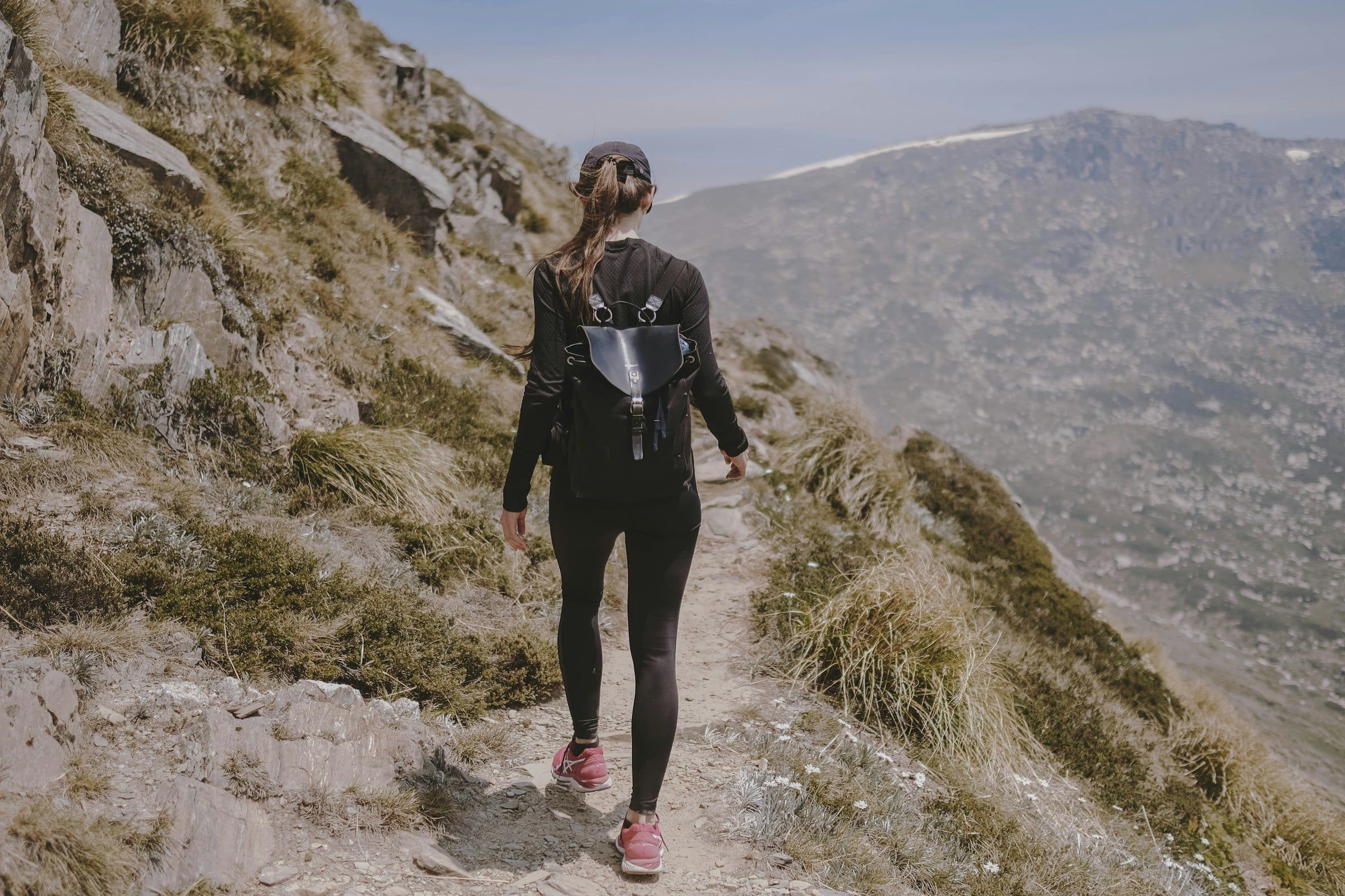 Escursionista di spalle con giacca tecnica nera e zainetto da trail mentre sale un sentiero roccioso di montagna