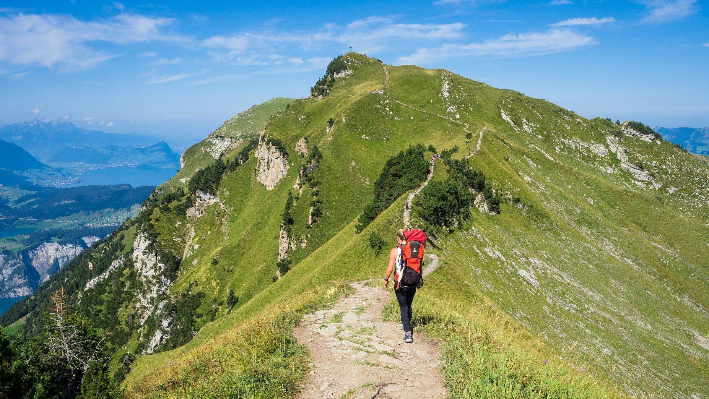 Escursionista con lo zaino rosso lungo un sentiero di cresta erboso, con un lago e le montagne che si aprono all'orizzonte
