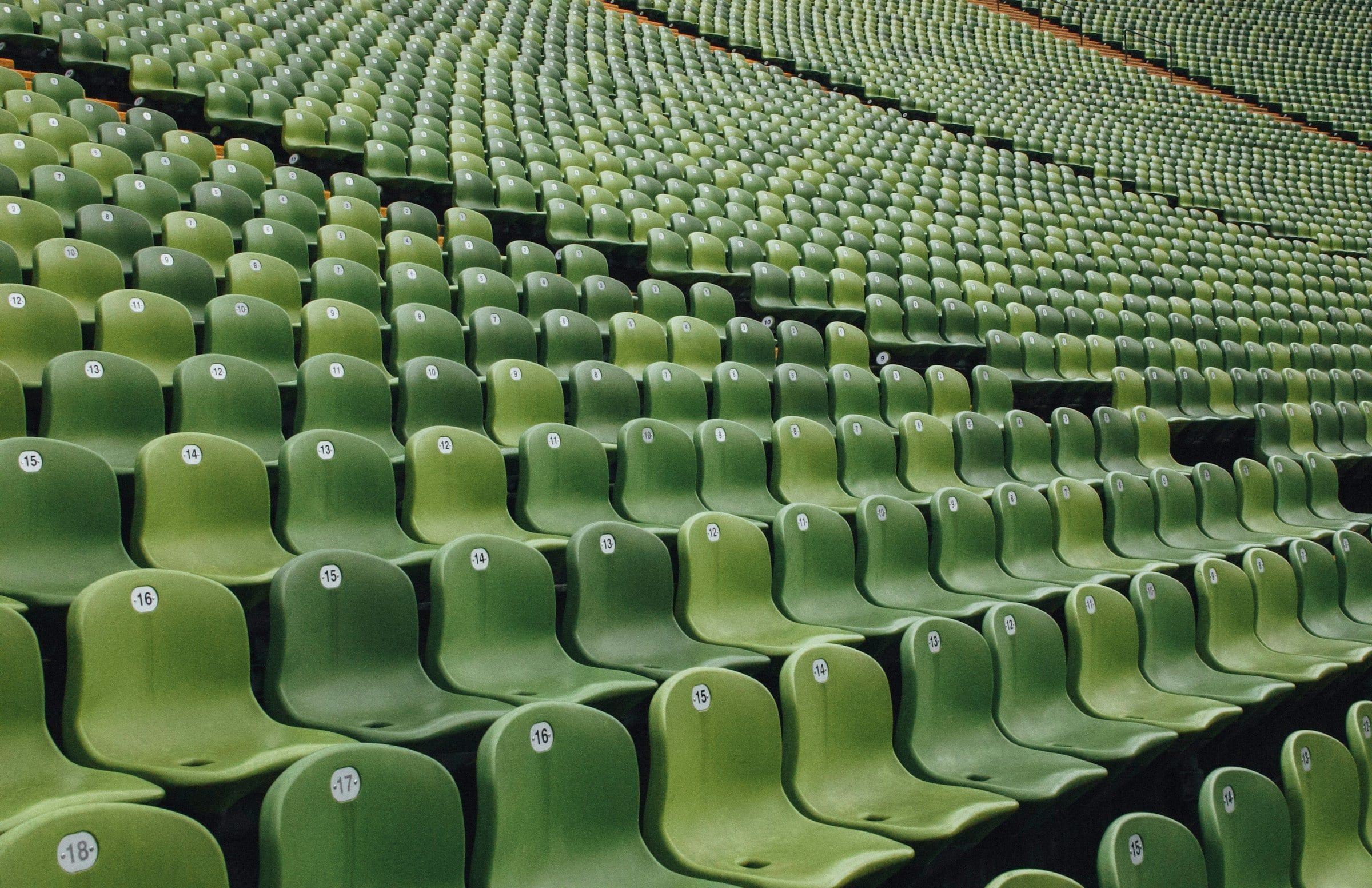 Gradinate di uno stadio con lunghe file di seggiolini verdi vuoti che salgono in diagonale, viste da vicino in piena luce