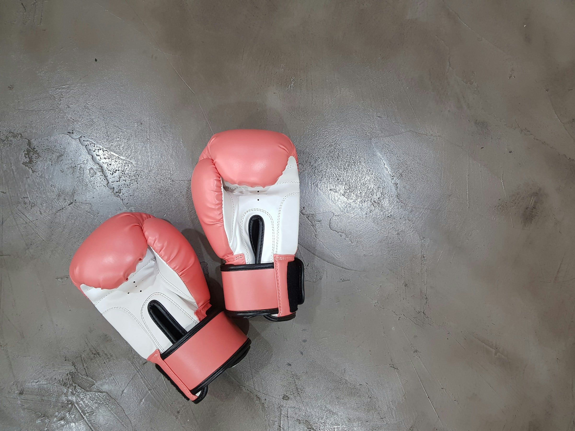 Due guantoni da boxe rosa e bianchi appoggiati su un pavimento di cemento grigio, ripresi dall'alto in una luce morbida