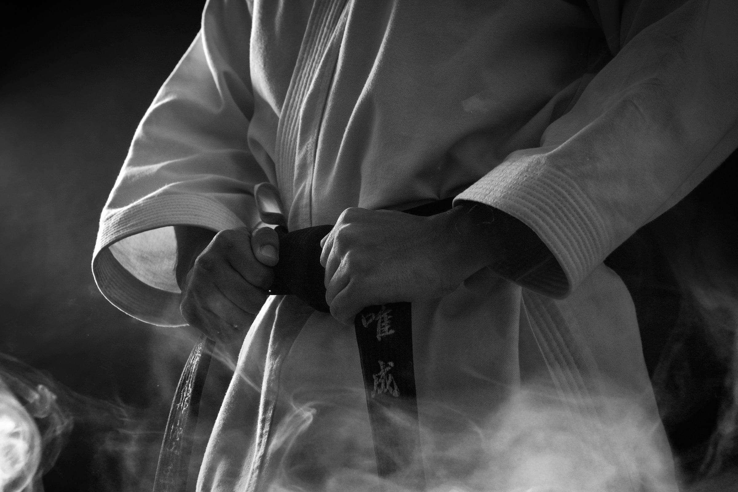 Primo piano in bianco e nero di un karateka in kimono bianco che stringe con le mani la cintura nera annodata in vita, avvolto da un leggero fumo