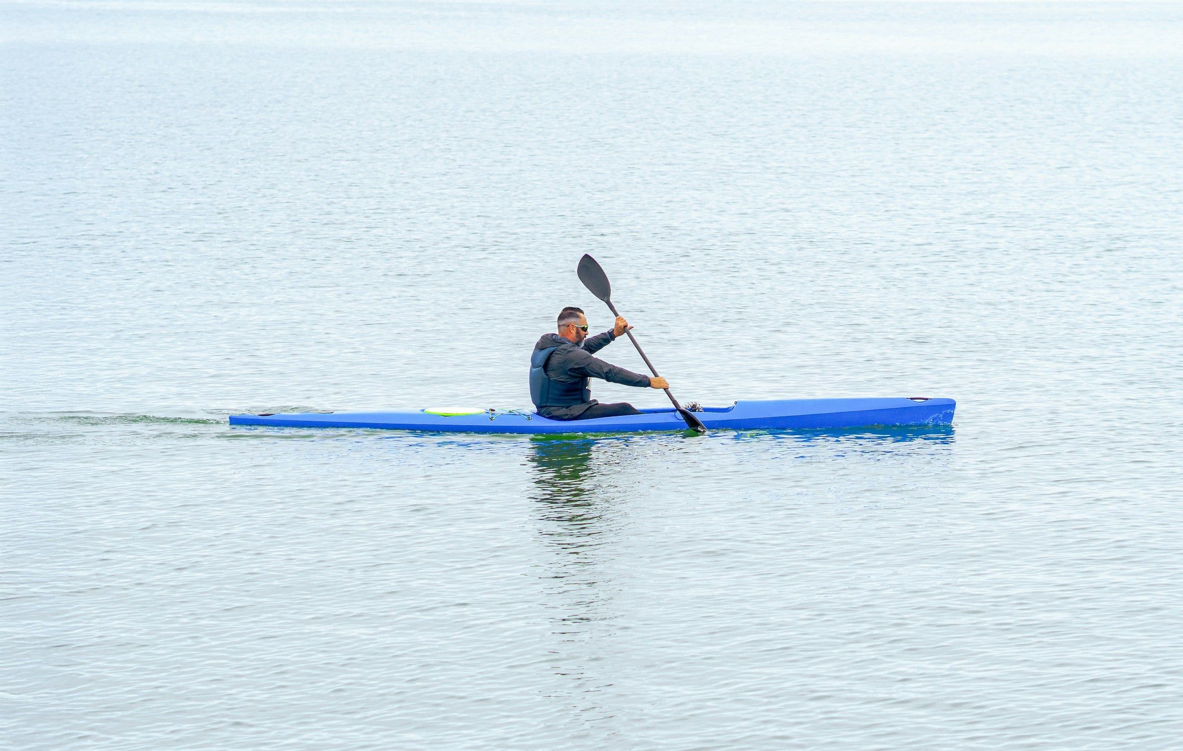 Canoista in kayak blu che pagaia su un lago calmo e grigio, unico punto di colore sull'acqua liscia fino all'orizzonte