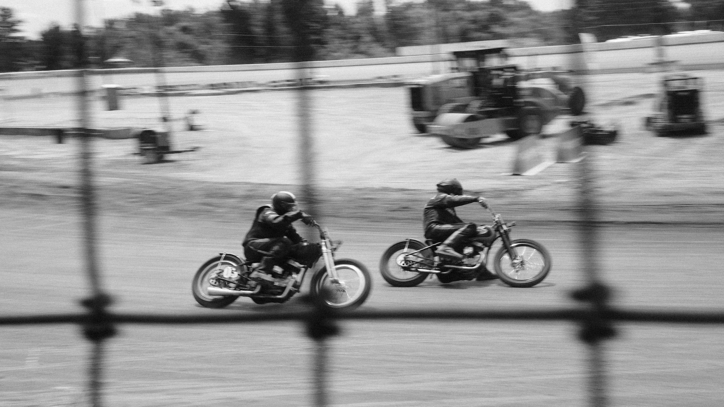 Due moto da corsa in derapata su una pista sterrata, fotografate in bianco e nero attraverso la rete della recinzione