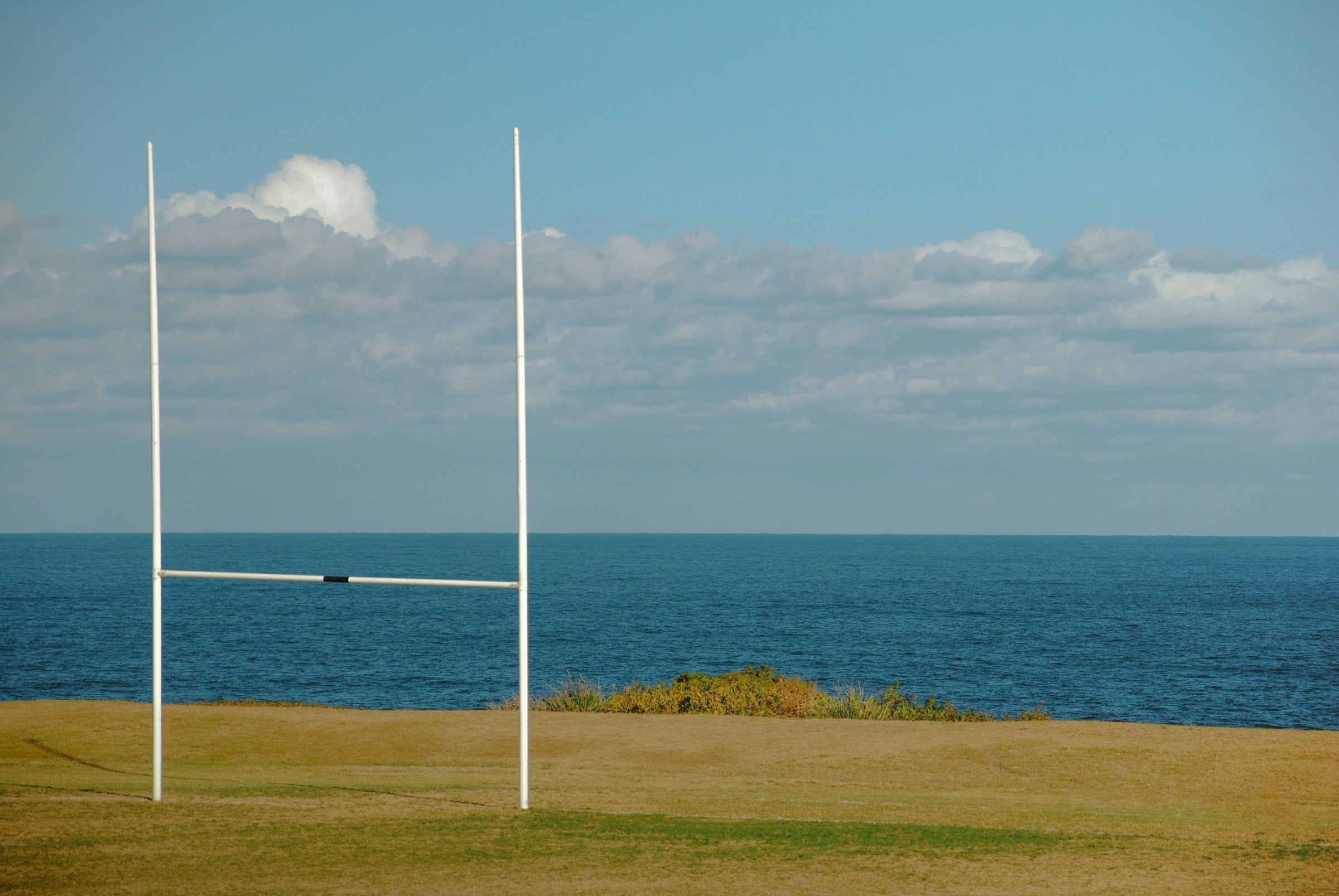 Pali da rugby a forma di H su un campo in erba affacciato sul mare, sotto un cielo azzurro attraversato da qualche nuvola bianca