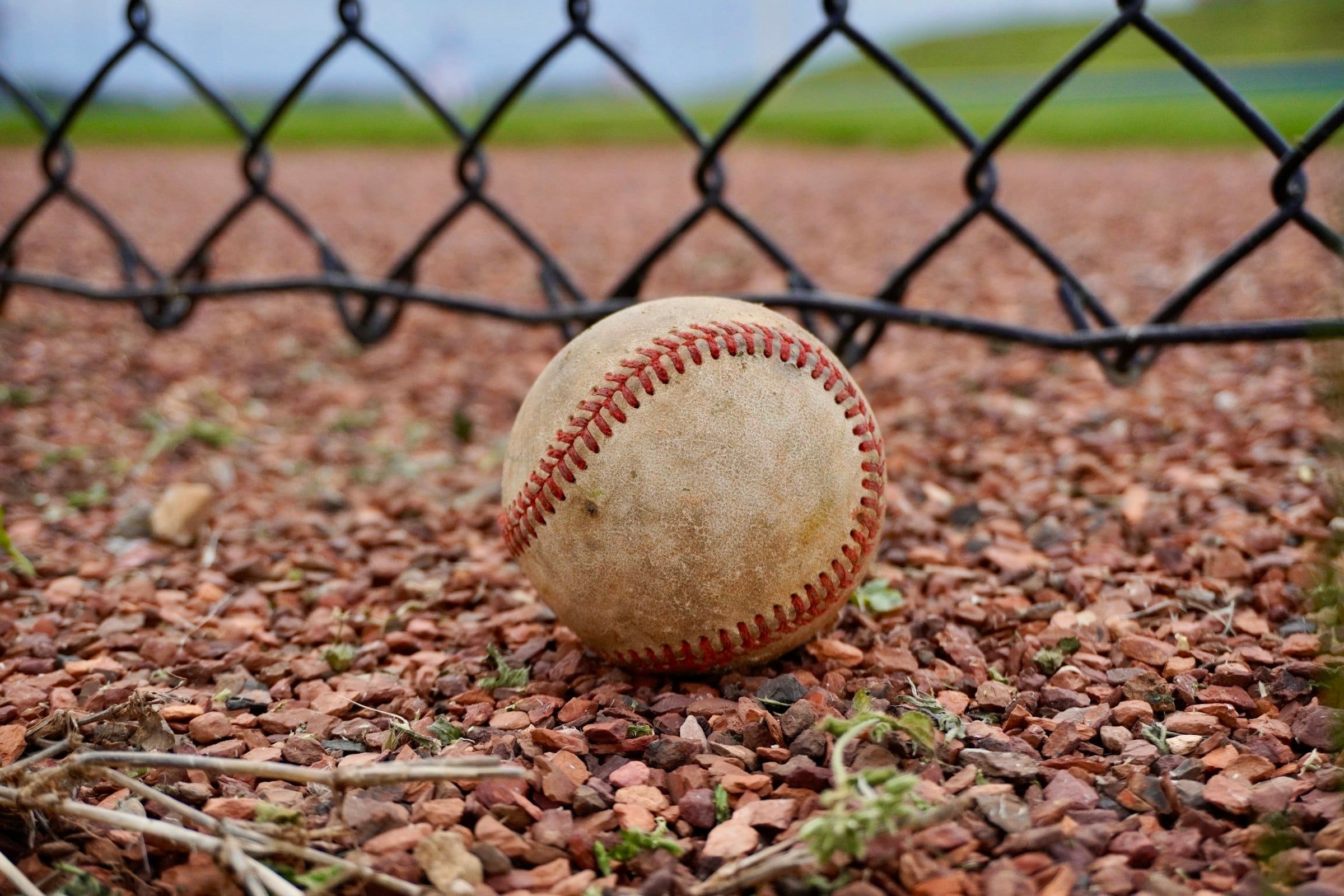 Palla da baseball consumata appoggiata sulla ghiaia rossa ai bordi di un campo, vicino alla base di una rete metallica