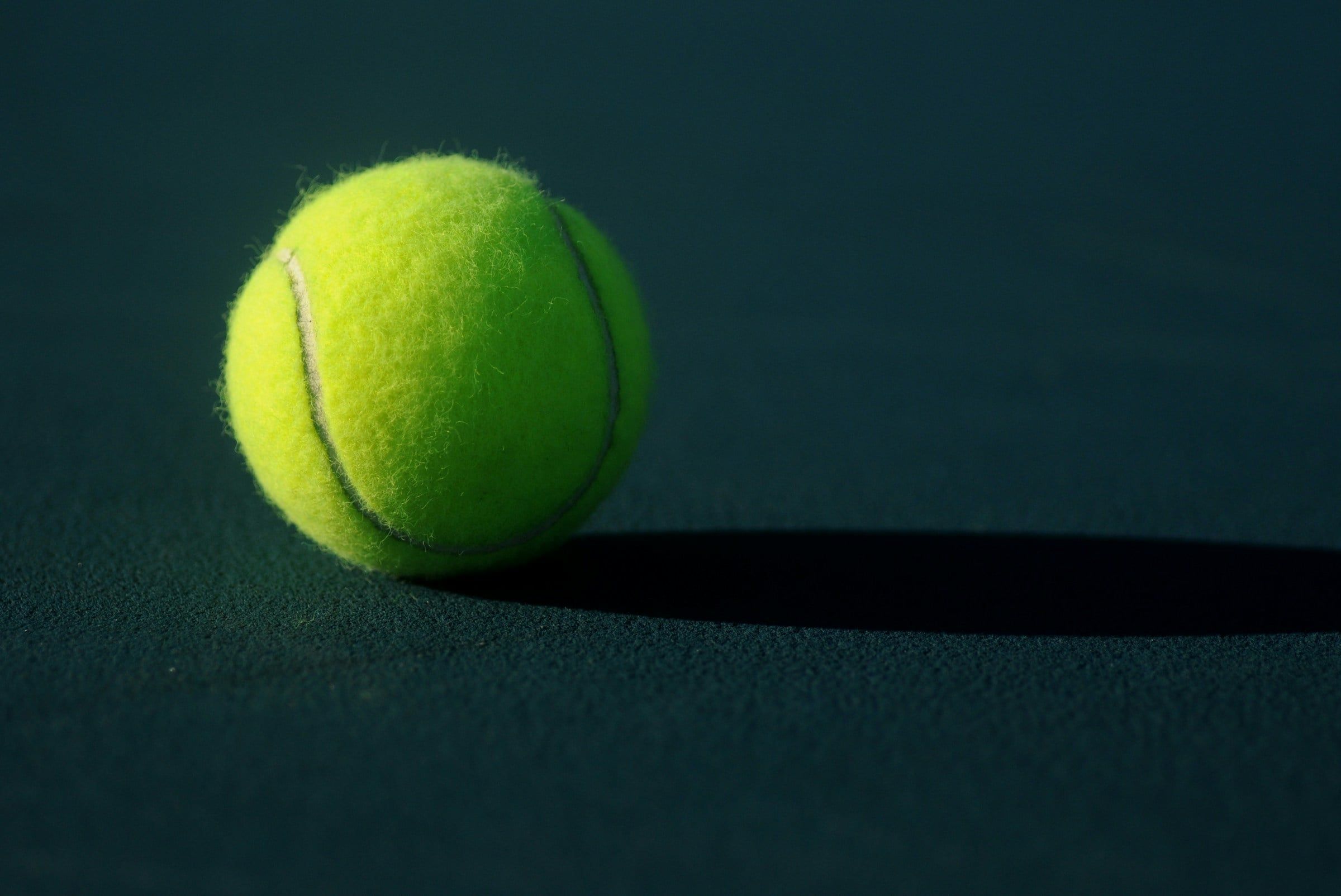Pallina da tennis gialla ferma su un campo dalla superficie scura, illuminata di lato con la sua ombra che si allunga accanto