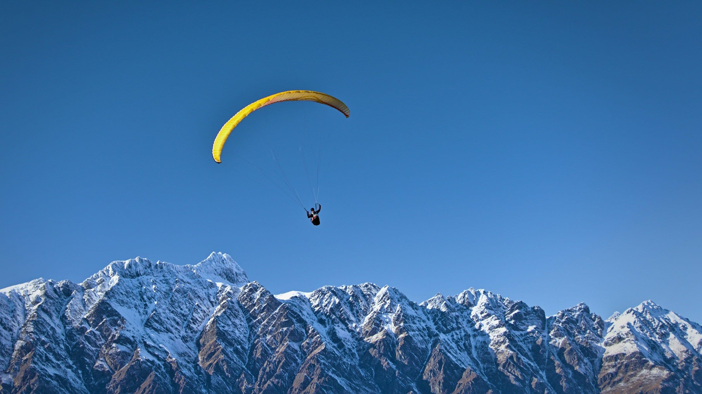Parapendio giallo in volo nel cielo azzurro sopra una catena di montagne con le cime coperte di neve