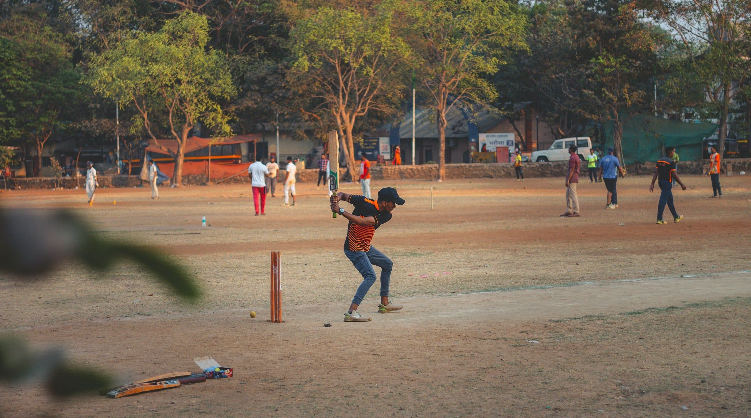 Partita di cricket su un campo di terra battuta all'ombra degli alberi, con il battitore davanti ai paletti e gli altri giocatori sparsi