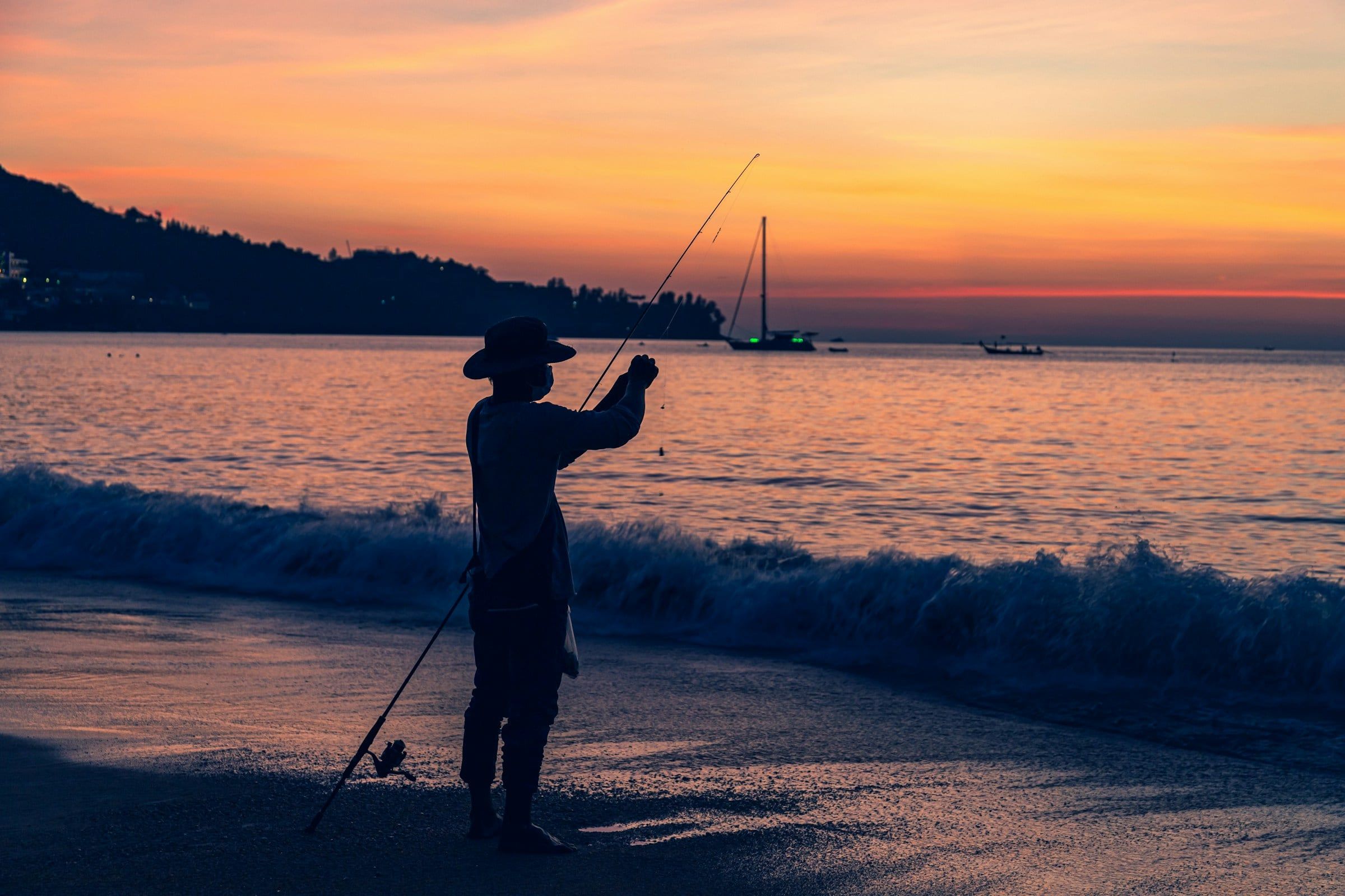 Pescatore in controluce sulla riva con il sole basso, con la canna da pesca protesa verso il mare calmo e il cielo arancione