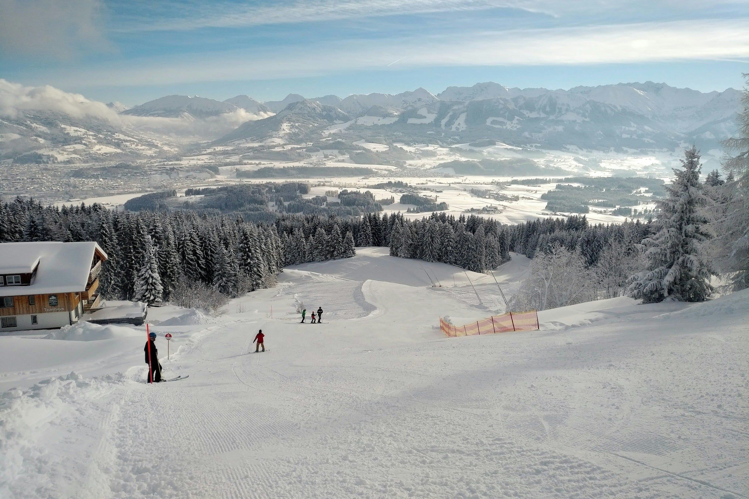 Pista da sci innevata che scende verso un bosco di abeti, con piccoli sciatori lontani, uno chalet in legno e le montagne all'orizzonte
