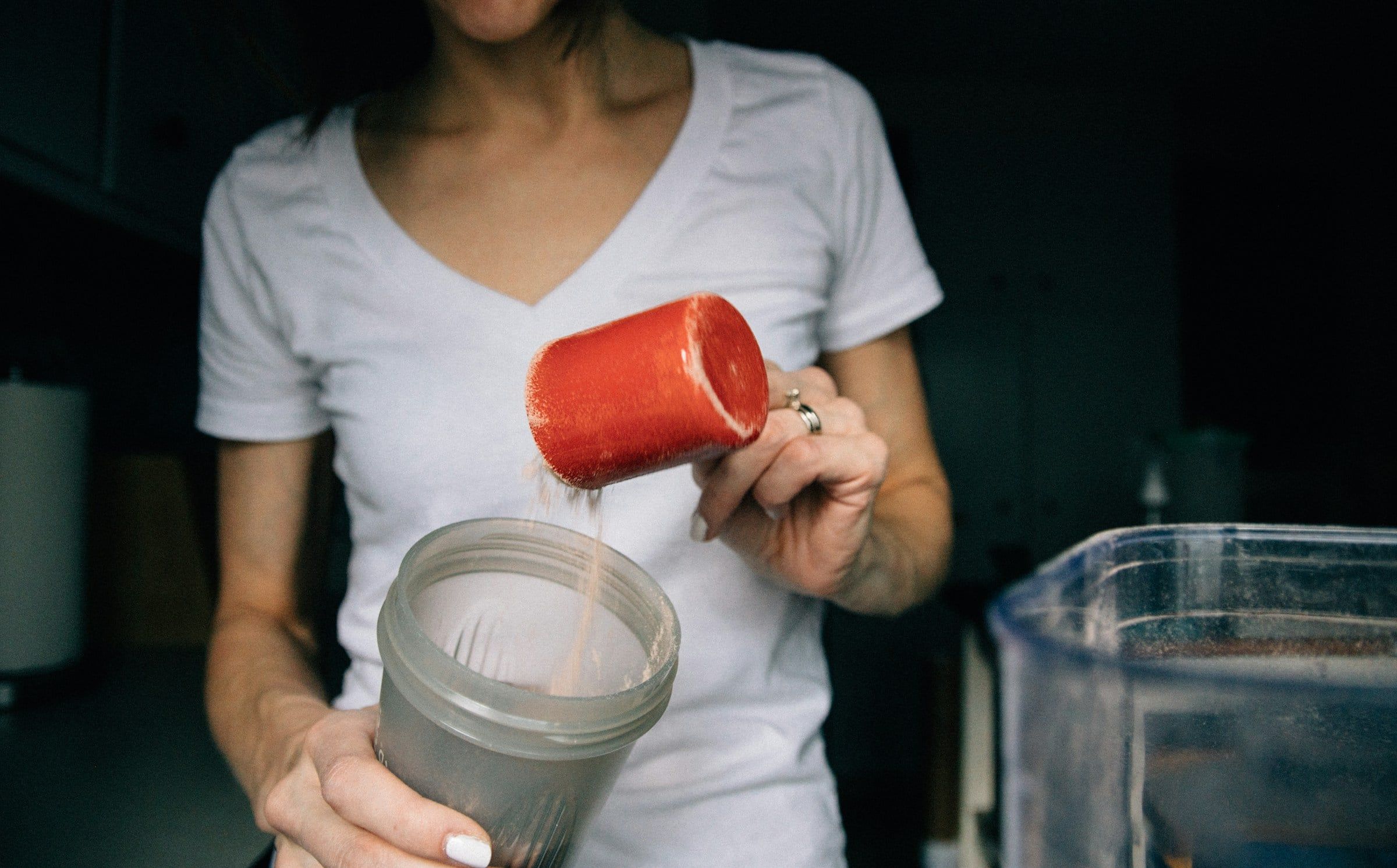Mani che versano la polvere da un misurino rosso dentro uno shaker trasparente, preparando un integratore accanto a una caraffa d'acqua