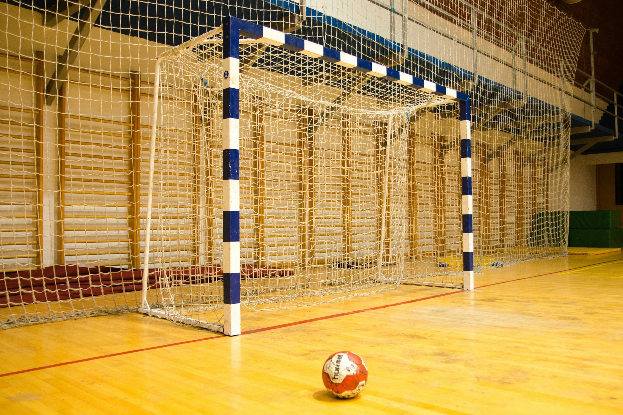 Porta da pallamano con la rete bianca in un palazzetto, con un pallone appoggiato sul parquet davanti all'area di porta