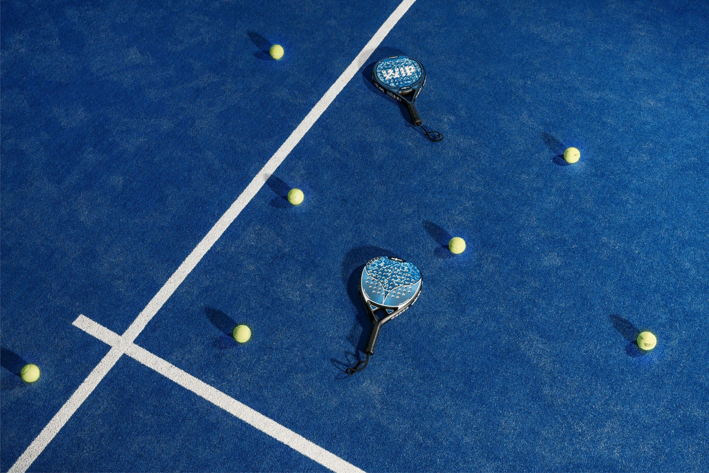 Due racchette da padel e alcune palline gialle sparse sul campo blu con le linee bianche, fotografate dall'alto