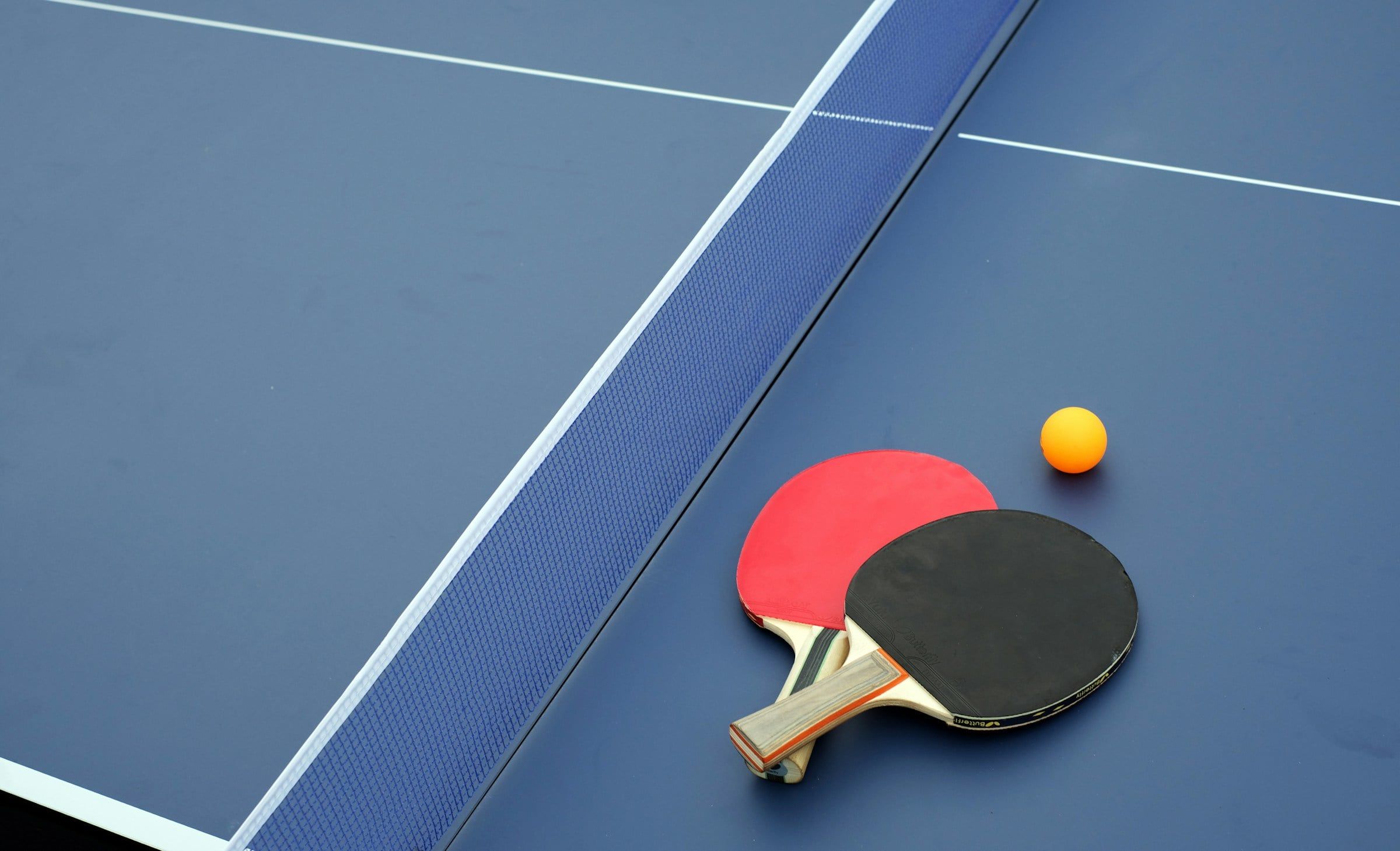 Due racchette da ping pong, una rossa e una nera, e una pallina arancione appoggiate sul tavolo blu con le linee bianche