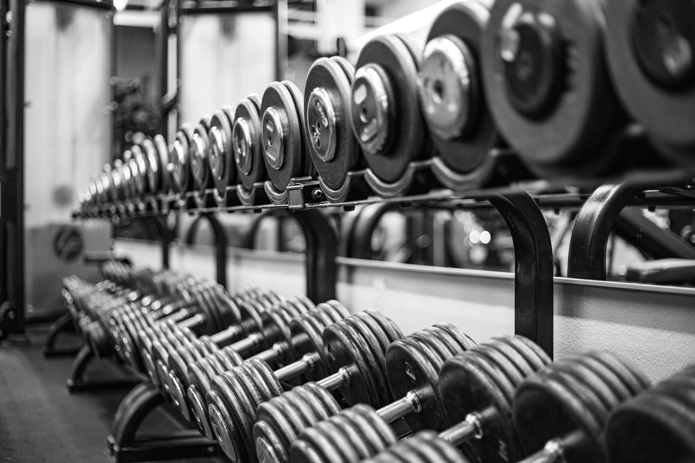 Rastrelliere di manubri in una palestra fotografate in bianco e nero, con le file di pesi allineate che si perdono in profondità