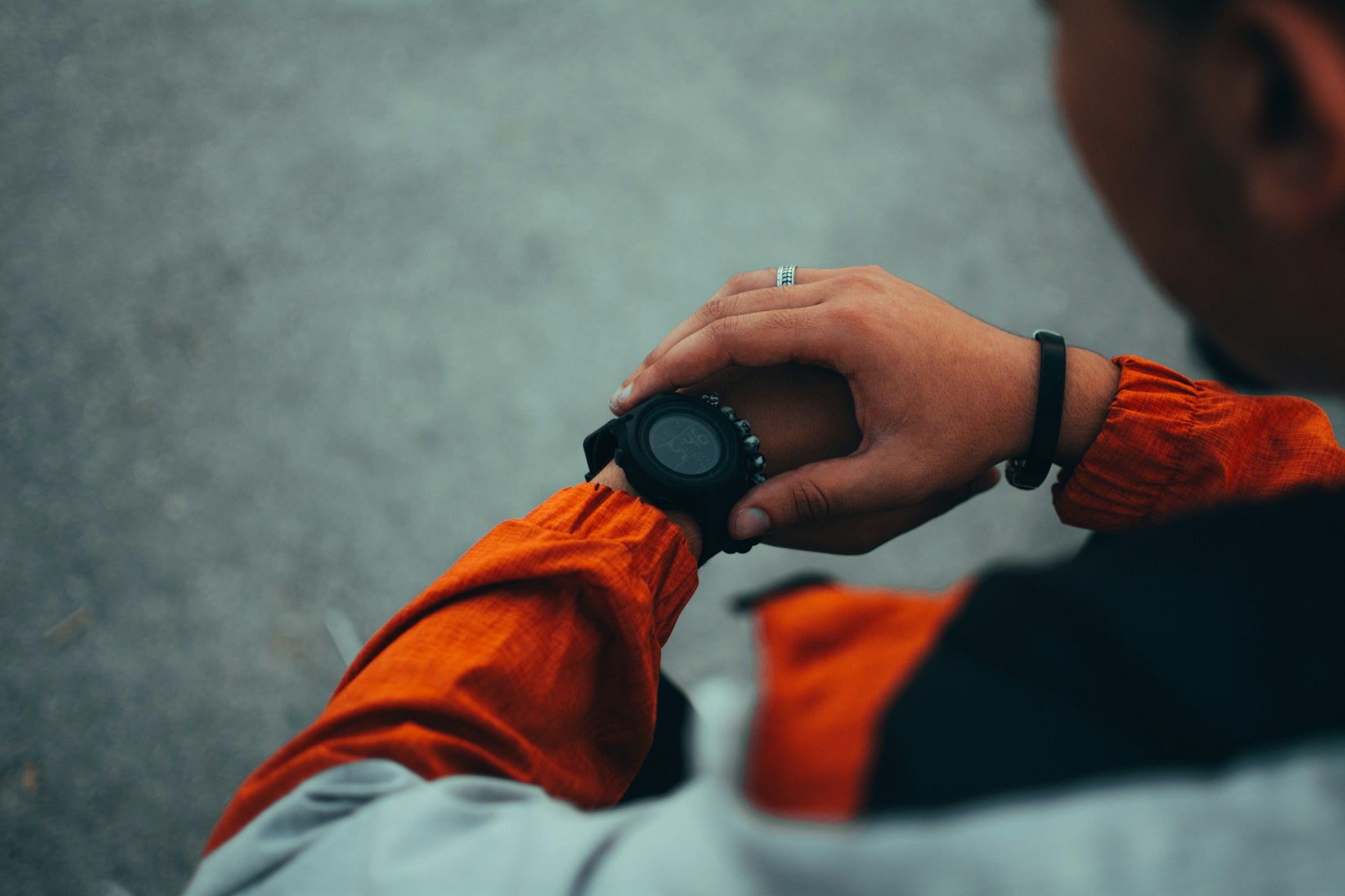 Runner con la giacca arancione ripreso di spalle mentre controlla l'orologio sportivo al polso durante una pausa all'aperto