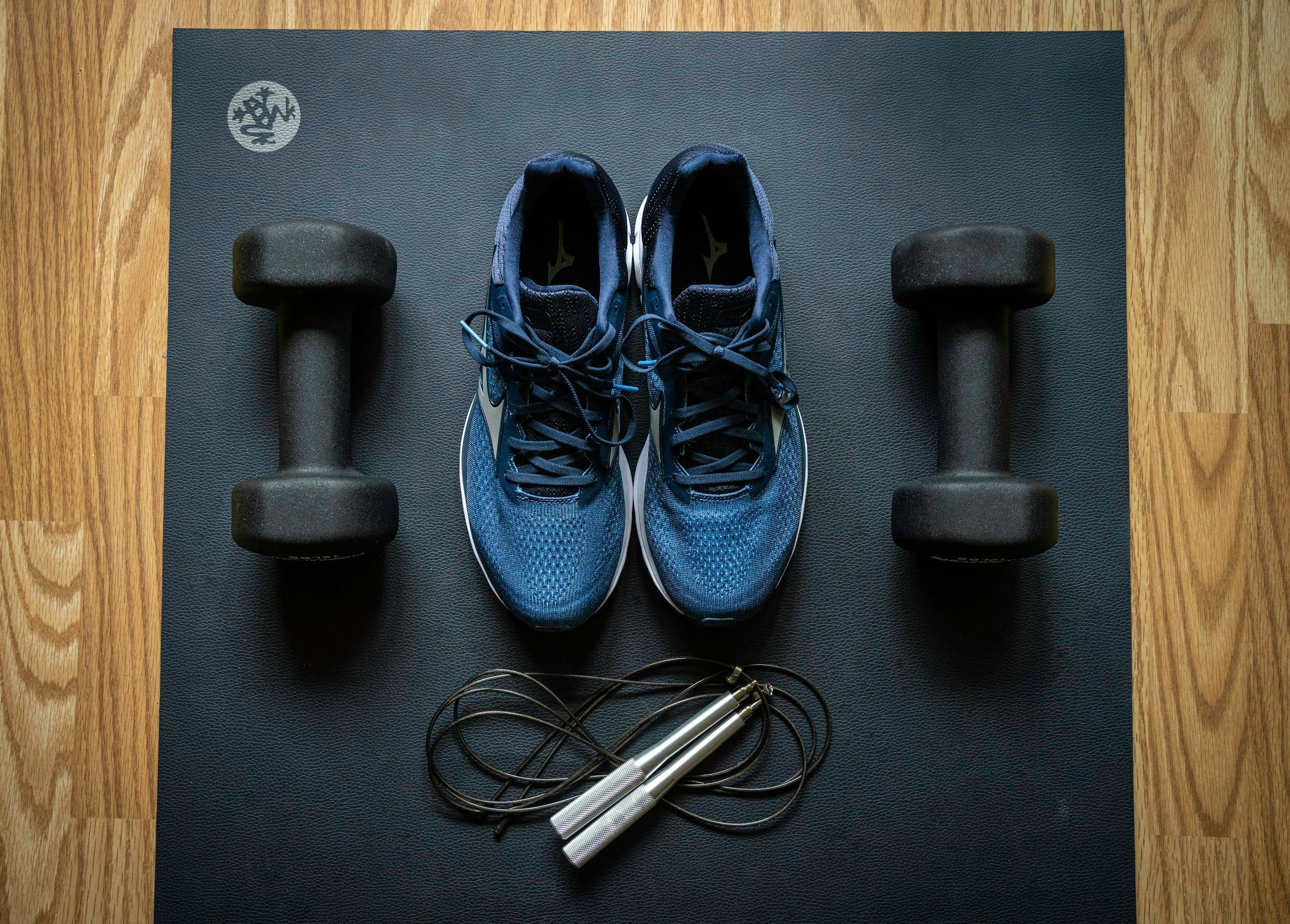 Scarpe da running blu tra due manubri neri e una corda per saltare, disposte in ordine su un tappetino scuro sul parquet