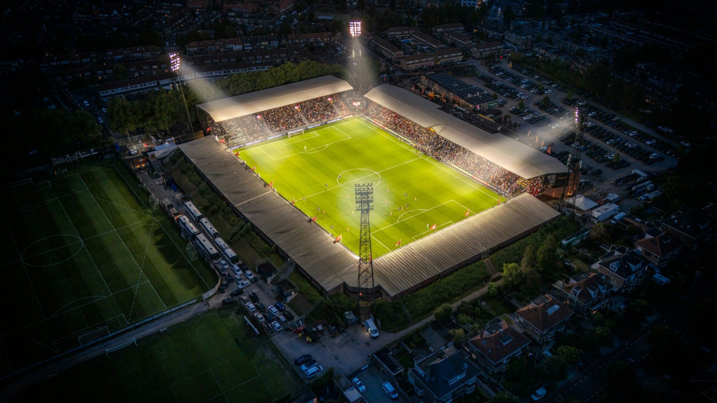 Vista aerea di uno stadio di calcio illuminato dai riflettori di notte, con il campo verde al centro e il quartiere buio tutto intorno