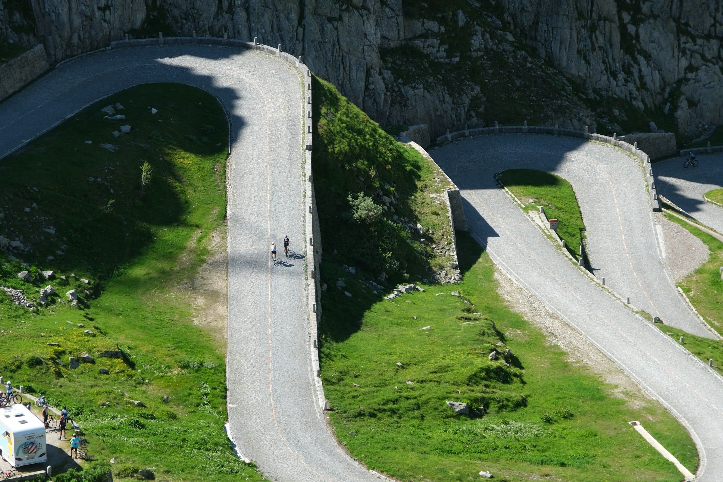 Tornanti di una strada di montagna tra prati e pareti di roccia, percorsi da alcuni ciclisti che salgono verso il passo